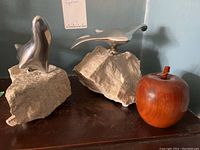 Two silver-tone metal figurines mounted on natural beige stone bases and one wooden apple sculpture placed on a dark wood surface under light.