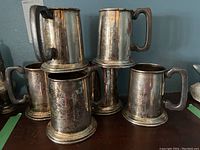 Six tarnished silver-tone metal beer steins arranged on wood surface against blue wall
