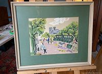 Full view of the framed painting showing the art market scene with green mat board and light-colored frame