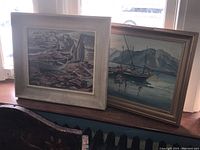 Both framed paintings shown together on a surface near window light.
