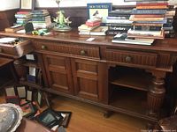 Front view of the wooden sideboard with books and assorted items on top, showing three drawers and double doors in center with open shelves on sides.