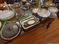 Wide view of assorted silver plate serving dishes, trays, fruit bowl, and Fire King casserole dish displayed on wooden table.
