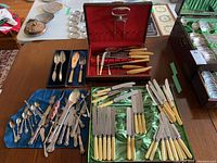 Photo showing various cutlery items spread on a table with the cutlery case open containing knives with cream-colored handles, and mixed silver plate forks and spoons.
