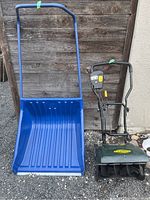 Yardworks snow thrower next to large blue Garant shovel on gravel surface against wooden wall.