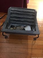 Electric tabletop water feature with ridged back panel and rectangular basin containing decorative stones, placed on a wooden floor.