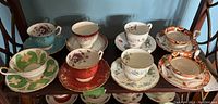 View of eight teacups with saucers of various floral and decorative designs including purple flowers, green floral swirl, red floral, and orange and black detail with gold trim.