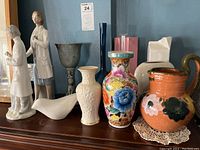 Overall view of various ceramic vases and figurines grouped together on a shelf including floral vases, jugs, bird figurine, and standing women figurines.