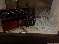 Wide view of eight assorted clear wine glasses beside two black wine bottle-shaped boxes holding metal wine opening tools and two decorative corkscrews in front.