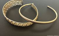 Two Kendra Scott Keeley gold tone cuff bracelets, one with detailed cutouts and one solid band, shown on black background