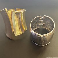 Two cuff bracelets shown together, one gold tone with smooth curved geometric design and one silver tone with hammered texture and hinge clasp.