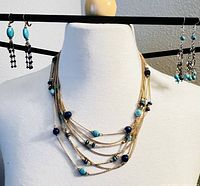 Layered necklace with turquoise and lapis lazuli beads displayed on a mannequin with two pairs of silver tone earrings with turquoise beads hanging on a metal stand.