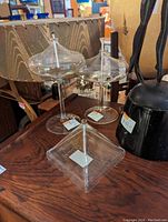 Photo showing three clear glass candle holders with tall stems and various shaped tops on a wooden surface, surrounded by other household items.