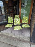 Four vintage chairs with green floral upholstery arranged outdoors on concrete.