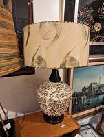 Photo of a retro style table lamp with beige fabric lampshade featuring brown swirl patterns. The base is ceramic with a speckled brown and white finish, mounted on a black stand.