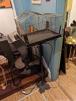 Bird cage with metal wire roof-shaped top on black metal stand in indoor setting.