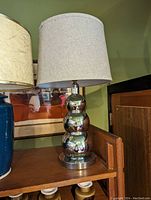 Chrome table lamp with three stacked spherical metal base and white fabric drum shade on wooden surface