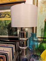 Full view of the chrome table lamp with white drum shade placed next to decorative glass bottles and framed artwork in the background.