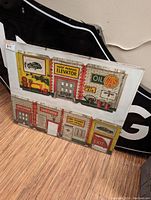 Framed art piece depicting vintage tin toy garage imagery with signs such as 'Roof Parking Elevator', 'Oil Gas', 'Car Wash', 'Exit', and 'Elevator', in a retro color scheme.