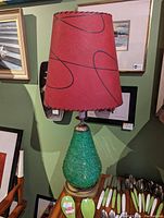 Full view of a table lamp showing a green textured resin base resembling spaghetti strands and a red tapered lampshade with black abstract lines, placed against a wall with framed artwork.