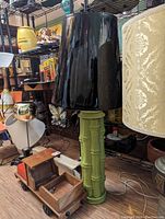 Photo of a green bamboo style lamp with a black shade placed on a floor with nearby shelving and other items.