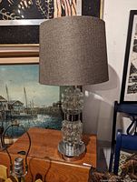 Full view of the glass base table lamp showing the bubble-textured clear glass base, metal bottom, and dark gray fabric drum shade.