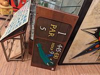 Photo of wooden golf score plaque with flip score numbers, par indicator, and an alligator design showing handicap numbers on a wooden wall.