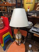Photo showing ceramic lamp with beige fabric lampshade, natural tree design on ceramic base, placed on orange chair among other vintage items