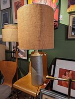 Photo of a large ceramic table lamp with beige linen shade, placed on a chair in a room with various framed artworks on the walls.