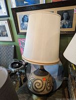 Large retro table lamp with rounded base decorated with abstract swirl and geometric patterns and a cream-colored fabric lampshade.