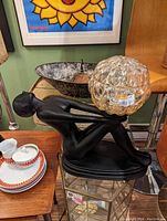 Photo of a black lamp depicting a reclining female figure holding a bubble-patterned amber glass globe lamp shade.