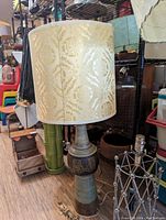 Mid-century ceramic table lamp with large cream and gold fabric shade standing on a shelf with other items in background.