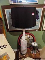 Tall table lamp with ribbed white ceramic base, brass accents, and large black cylindrical lampshade on wooden surface.