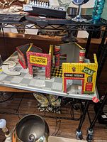 Photo of multiple vintage tin garage playset pieces on a glass shelf, showing entrance, elevator, and gas/oil signs, along with base platform and pillars.