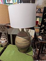 Photo of a ceramic table lamp with a white drum lampshade on a green surface, showing the rounded striped ceramic base.