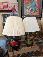 Photo showing two Gilbert table lamps with ceramic bases and white pleated lampshades. One base is dark red and black, other base is beige, brown and mustard yellow.
