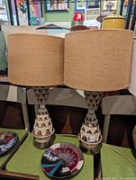 Photo showing a pair of retro table lamps with beige fabric drum shades and sculpted ceramic bases with brown and cream geometric patterns on brass-colored stands.