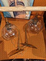 Photo showing two brass wall light fixtures with elegant curved arms and clear glass globe shades lying on an orange fabric surface. An unopened rolled item rests above the lights.