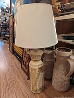 Photo of a plaster lamp with a beige fabric lampshade sitting on the floor in a room with other decor items.