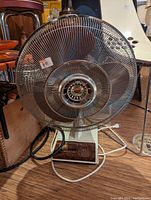 Close-up photo of retro metal electric fan with circular cage and base controls on wooden floor.