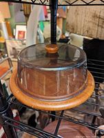 Clear image showing a round teak cheese board with glass dome cover on a metal rack, highlighting the teak knob on top and the natural wood grain of the board.