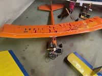Orange model airplane with damaged and torn wing covering, visible on floor with wing detached.