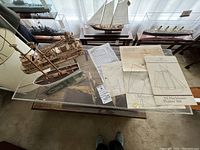 Two small wooden ships and related blueprints under glass on a table.