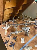 Overview of model airplanes arranged on wooden surface under stairs, showing multiple military aircraft models in different colors and designs.
