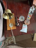 Brass bell mounted on wood plaque, carved pipe, glass paperweight, and glass thermometer on wooden base on a wooden surface.