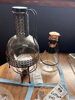 Photo showing two vintage carafes on a wooden table; one large ornate glass carafe with black metal latticework holder and candle warmer, and one smaller clear glass carafe with wooden stopper.