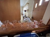 Photo showing six pieces of pink Depression glass consisting of four dessert/sherbert dishes and two candleholders arranged on a wooden shelf.