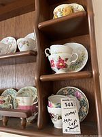 Shelving unit displaying multiple vintage teacups and saucers with varied floral designs, including yellow, pink, and purple flowers. Cups and saucers are arranged in pairs on wooden shelves.