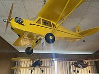 Full side view of the yellow model airplane hanging against ceiling tiles, showing propeller, wings and wheels.