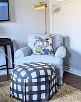 Single buffalo check ottoman placed in front of armchair with pillow and floor lamp, showcasing pattern and size.