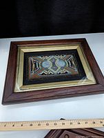 Framed piece showing a wooden and gold-tone frame enclosing the framed beadwork and medals with a measuring ruler below.